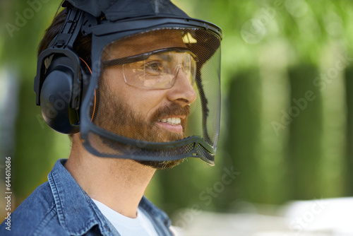 Close up view of bearded craftsman in protective face shield preparing for hard work outdoors. Bearded caucasian handyman in earmuffs looking ahead, with blurred background. Concept of protection.