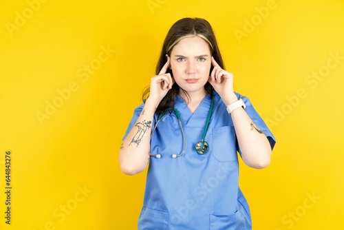 Serious concentrated Young caucasian doctor woman wearing blue medical uniform keeps fingers on temples, tries to ease tension, gather with thoughts and remember important information for exam