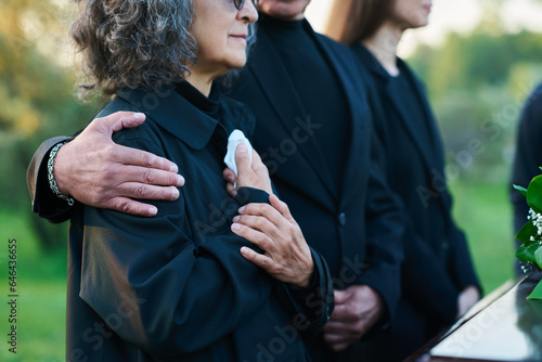 Hand of mature man on shoulder of his wife or sister with handkerchief lamenting passes away relative or family member during funeral service