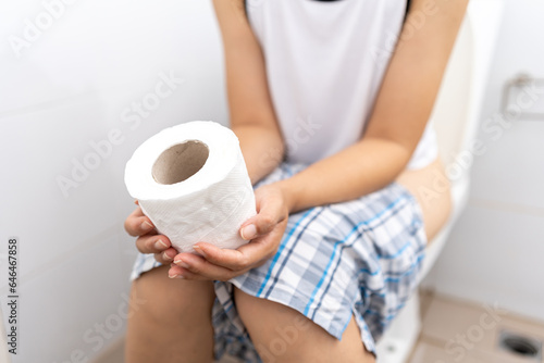 Female holding toilet paper roll and sitting on toilet in restroom. Woman squeezing tissue with diarrhea excreting in toilet. Hand holding a roll toilet paper is feeling pain with constipation.