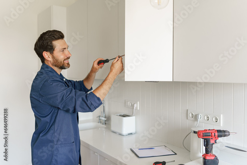 Professional repairman in uniform is using a screwdriver to adjust door of cabinet in kitchen