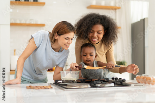 Multiracial lesbian and married girlfriend standing together at kitchen counter playing with their biracial son. Gay women living affectionate life parenting their kid. LGBT, homosexual relationship.