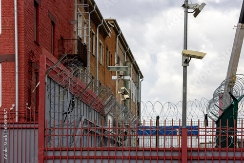 barb wire and surveillance cameras over fence in city near red brick building wall at summer day, telephoto closeup with selective focus