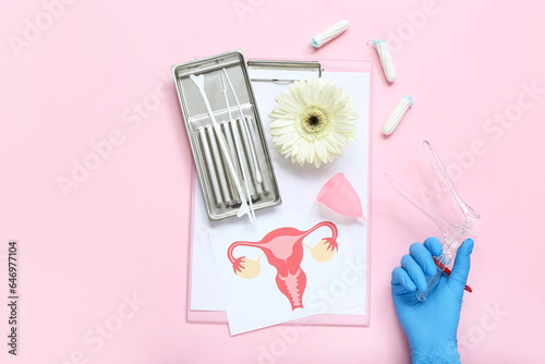 Hand in medical glove, with gynecological speculum, pap smear test tools, drawing of female uterus, menstrual cup and tampons on pink background