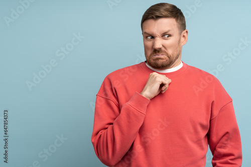 Confused man feeling disgust with serious face incredulously looking aside isolated on blue background. Clenching fist gesture on chest. Guy has scared expression, demonstrates aversion makes grimace.