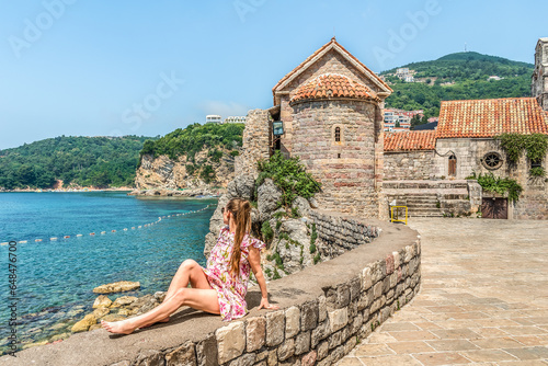 A young woman poses while sitting on a parapet in the Old Town of Budva, against the backdrop of layered rocks and the Adriatic Sea. View from the ancient fortress with a stone tower to Mogren beach