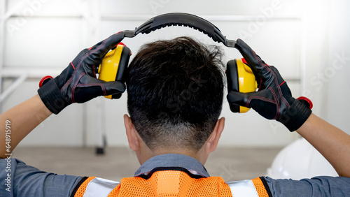 Back view of male worker with reflective orange vest and protective hand gloves pulling yellow safety ear muffs or ear protectors when wearing on his head. Equipment for high noise reduction