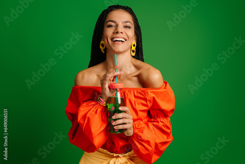 Joyful young woman holding bottle with lemonade and looking at camera against green background