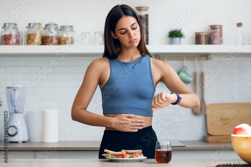 Hungry sporty woman waiting for the time to eat healthy breakfast after fasting in the kitchen at home