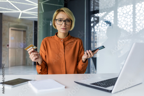 Portrait of disappointed and deceived woman inside office at workplace, businesswoman holding bank credit card and phone, looking upset at camera, received error and rejected money transfer