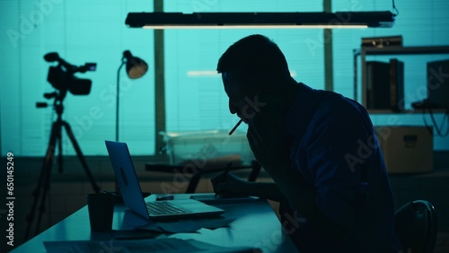 Medium silhouette shot of a detective, policeman sitting in the interrogation room, smoking, working on a laptop and studying the evidence.