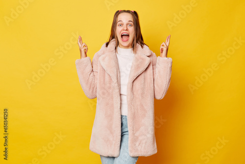 Pretty brown haired girl wearing pink fur coat with ponytails isolated over yellow background stands with crossed fingers warm smile emanating strong belief in positive outcomes