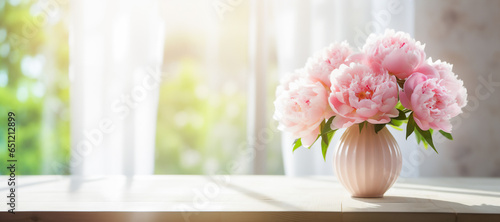 A vase with ribbed texture of light pink peonies on a wooden table in front of a window with white curtains. The background is a garden or park with green trees and grass. Bright and airy mood.