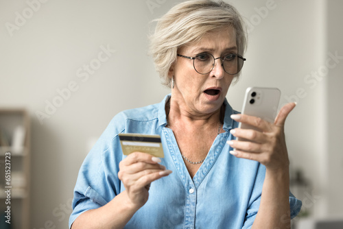 Shocked concerned mature woman in glasses holding credit card, staring at smartphone in bad surprise, getting stress, financial problems, money stealing, overspending, bankruptcy risk