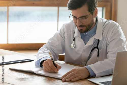Focused doctor wearing glasses writing illness history, sitting at workplace in hospital, serious man gp therapist wearing white coat uniform with stethoscope filling patient card, paperwork