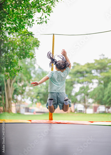 Asian child girl is jumping on trampoline on playground background. Happy laughing kid outdoors in the yard on summer vacation. Jump high on trampoline. Activity children in the kindergarten school.