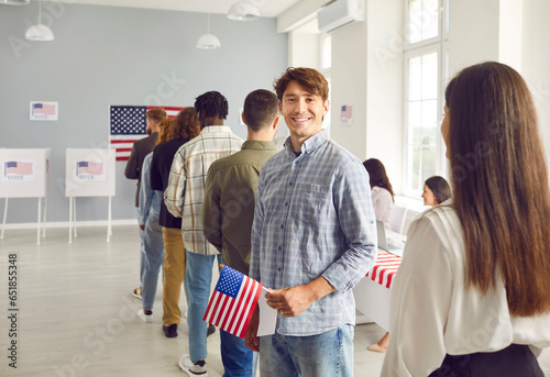 Happy male American voter comes to vote at polling station. Young man with flag of USA in hand standing in line with other people, looking at camera and smiling. US elections concept