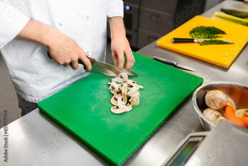 cooking food, profession and people concept - close up of male chef with knife chopping champignons on cutting board at restaurant kitchen