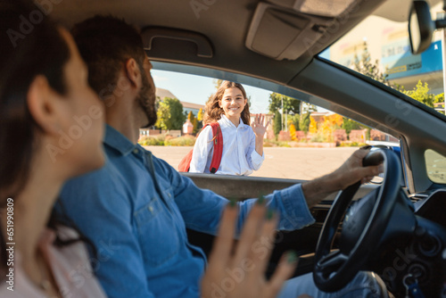 Happy parents bringing their daughter to school, sitting in car and waving hands to child girl