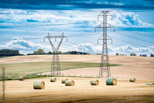 Transmission towers and power lines overlooking a farm field during fall harvest with round hay bales on the Canadian prairies in Rocky View County Alberta Canada.