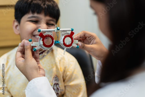 Happy Indian child boy with women eye specialist examining eyesight modern ophthalmology equipment in clinic. Patient kid male checkup iris examines ophthalmological hospital. measure eyeglasses.