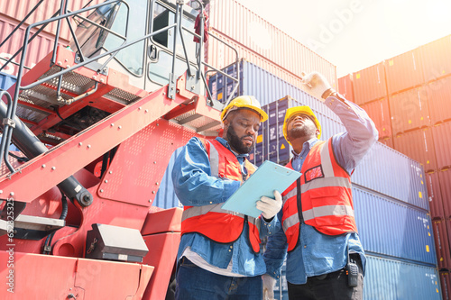 Two African American logistic workers wearing reflective vests and white helmets talk about logistics operations at shipping container yard. Transportation import and export logistic industry concept.
