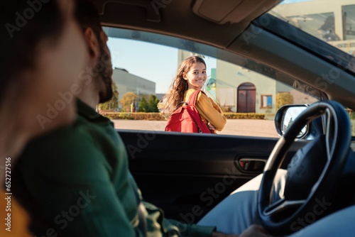 Mother and father dropping off daughter in front of school, girl turning back to parents sitting in car and smiling