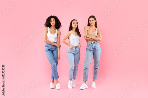 Three serious diverse women posing standing over pink background, full length shot of ladies trio looking at camera