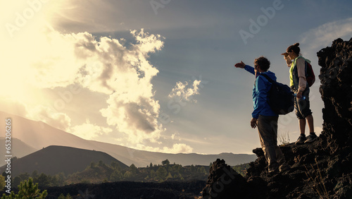 Hiking Couple Admiring Vast Landscape: A mature couple in windbreakers and with backpacks stand atop a rock, taking in the view while shielding their eyes, with an expansive sky perfect for copy.