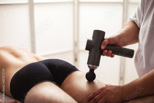 Doctor therapist treating injury of leg muscle sportsman of gun percussion massager. Masseur doing massage of thigh guy in medical clinic room. Alternative therapy rehabilitation. Copy ad text space