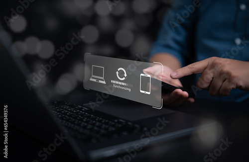 Mobile phone synchronize data with a computer for backup over a wireless connection. Hand-holding smartphone to sync sharing files or information to laptop with virtual progress update screen.