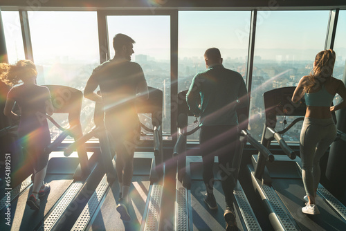 Rear view of athletic people running on treadmills in a gym with city view. Silhouettes of group of four people doing cardio in modern indoor environment.