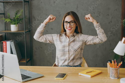 Young fitness employee business woman wear shirt casual clothes glasses sit work at office desk with pc laptop show biceps muscles on hand demonstrating strength power. Achievement career job concept.