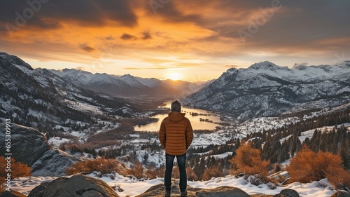 Person standing on a snow-covered ledge overlooking the stunning sunset vista of mountains.