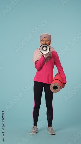 Full-sized isolated photo capturing an attractive young woman wearing a sportswear and a hijab, sheila, holding an exercising mat. She is actively speaking in a rupor, loudspeaker.