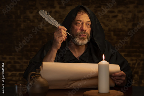 An old monk with a beard holds a quill in his hand over a parchment against a dark brick wall, dark tonality.