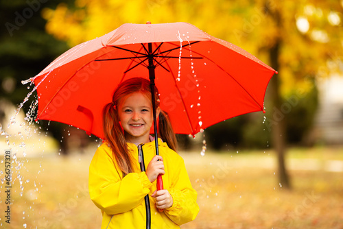 Happy child under summer rain. Funny girl have fun time while playing outdoors under autumn shower. Kid with umbrella and waterproof yellow boots.