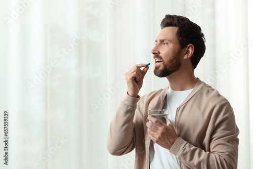 Handsome man with glass of water taking pill indoors, space for text