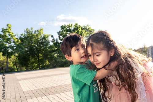 Little Brother Telling A Secret To Her Older Sister In A Park While Hugging Her At Sunset. She Is Listening Paying Attention.JPG