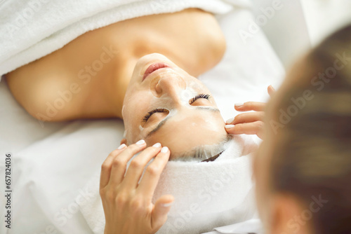 Professional spa therapist beautician applies white rejuvenating anti age lifting peeling antioxidant detox vitamin sheet mask on face of happy pretty attractive woman for soft smooth good complexion