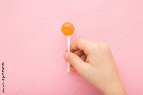 Little child fingers holding orange lollipop on stick on light pink table background. Pastel color. Sweet candy. Closeup. Top down view.