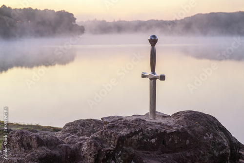 Excalibur l'épée légendaire plantée sur son rocher devant un étang dans la brume du matin au lever du soleil en forêt de Brocéliande