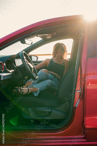 Pretty woman with sunglasses is posing inside of a car at sunset