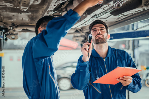 Car repair service. Two handsome young technician checking car condition in garage, Automotive mechanic pointing flashlight following at maintenance checklist document. Professional teamwork.