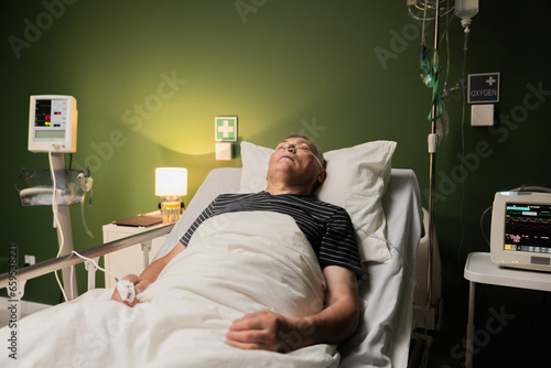 Elderly man in a hospital room, using an oxygen tube for respiratory support, within a ward