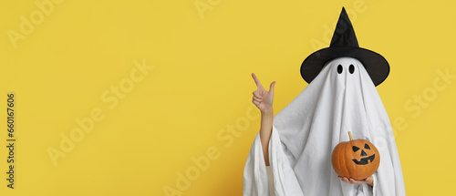 Person in costume of ghost holding Halloween pumpkin and pointing at something on yellow background with space for text