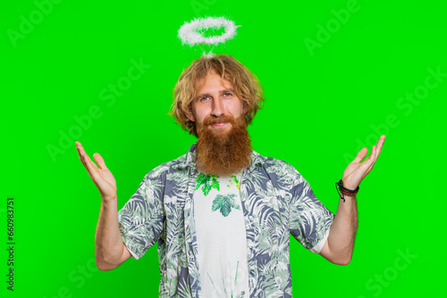 Portrait of smiling shy angelic young caucasian man with angel halo nimb over head flirting, looking at camera, positive love emotions celebrating holiday. Redhead bearded guy on chroma key background