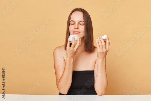 Satisfied young woman wearing black top holding container with moisturizer facial cream isolated over beige background smelling pleasant aromat of skin care product.