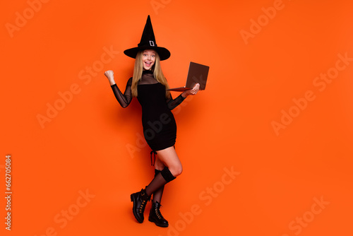 Full length photo of lovely blonde teen lady netbook raise fist luck dressed black halloween garment isolated on orange color background