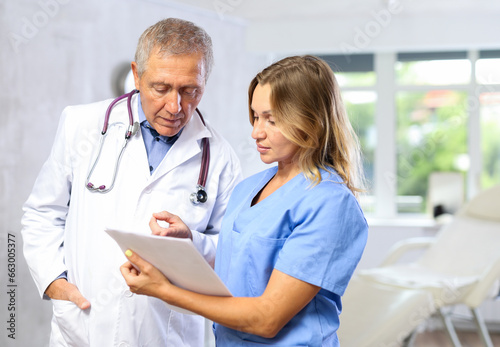 Two fellow doctors, adult woman and senior man, standing in medical office with papers in hands, focused on discussing of clinical diagnosis of patient